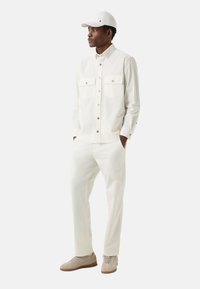 Biała koszula zapinana na guziki z dwoma kieszeniami, kremowe spodnie i beżowe buty. Do tego biała czapka, prezentująca minimalistyczny design.