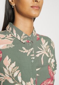 Camicia col colletto in tessuto verde con motivi floreali e farfalle rosa. Presenta una chiusura frontale con bottoni coordinati e maniche corte.