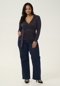 Top oscuro púrpura con brillo, con mangas largas y un lazo en la cintura, combinado con jeans rectos de mezclilla oscura y tacones negros.
