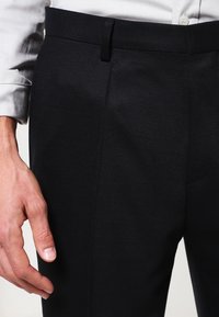 Svarta texturerade byxor med rak passform, som har en platt framsida, bältesömmar och en subtil lyster. Närbild med en hand som vilar i närheten.