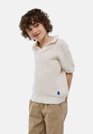 Pull polo en tricot crème à manches courtes, avec un col en V et un accent de logo d'arbre bleu près de l'ourlet. Associé à un pantalon marron clair.
