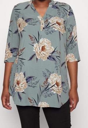 Kobieta ubrana w zieloną kwiecistą tunikę z beżowymi i fioletowymi kwiatami oraz rękawami do łokcia, w połączeniu z czarnymi spodniami.