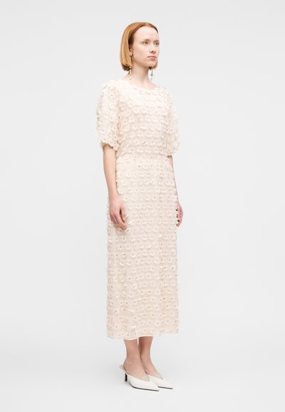 Robe midi beige avec appliqués floraux texturés, manches courtes bouffantes et silhouette droite. Associée à des talons blancs à bout pointu.