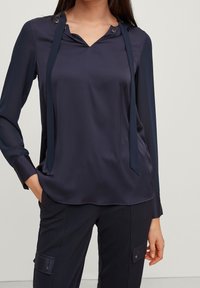 Navy blue blouse with a v-neck, long sleeves, and tie accents. Features a smooth, glossy texture and metallic buttons near the neckline.