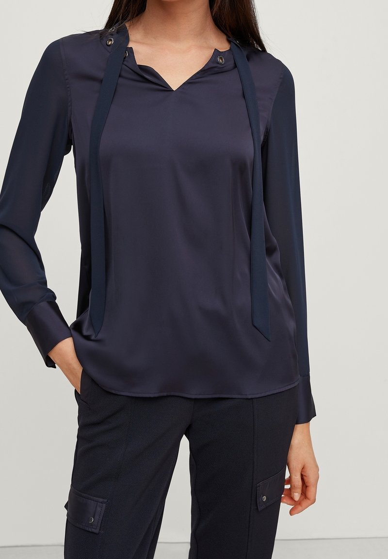 Navy blue blouse with a v-neck, long sleeves, and tie accents. Features a smooth, glossy texture and metallic buttons near the neckline.