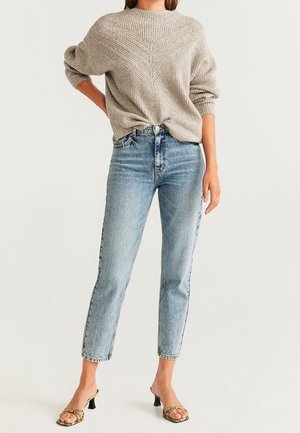 Frau trägt beigen Strickpullover, hellblaue Jeans mit hoher Taille und beige offene Sandaletten mit Absatz vor einfarbigem Hintergrund.