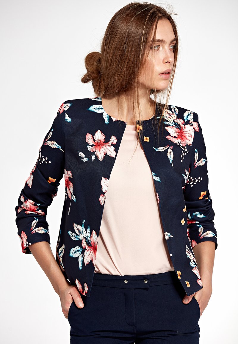 Kobieta z jasnobrązowymi włosami związanymi z tyłu, ubrana w granatową marynarkę w kwiaty, pod którą ma jasnoróżową bluzkę i granatowe spodnie, z rękami w kieszeniach, patrzy w prawo.