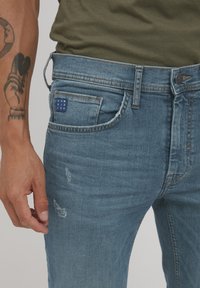 Jeans de mezclilla azul desgastados con áreas desgastadas y un pequeño parche de la marca azul en el bolsillo trasero. Presenta un diseño estándar de cinco bolsillos.