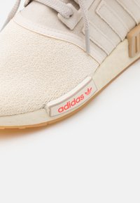 Beige textilsneaker med ljusgrå detaljer, med gummisula och en orange adidas-logotyp på sidan. Snörena är ljusgrå.