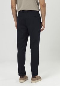 Marineblauwe slim-fit broeken met een gladde textuur, uitgevoerd met een discreet tailleband en twee achterzakken. Gecombineerd met lichtbruine sneakers.