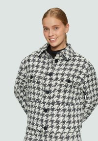 Een jasje met houndstooth-print in zwart en wit, met een kraag, voorzakken en zwarte knopen op een gestructureerde stof.