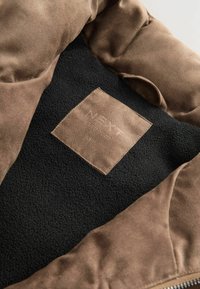 Unutrašnjost smešnog antilop kaputa u smešnoj boji sa crnim flis podstavljanjem i smešnom etiketom s natpisom "NEXT" ušivenom. Vidljiv je ivica sa rajsferšlusom sa strane.