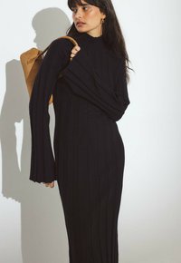 Ženska z dolgimi temnimi lasmi, oblečena v črno pleteno obleko, ki nosi rjav rjavnik iz semiša, gleda na stran proti enobarvni podlagi.