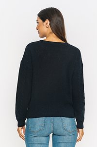 Navyfarbener Strickpullover mit lässigem Schnitt, Kabelstrickärmeln und rundem Ausschnitt; kombiniert mit hellblauen Jeans.