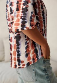 Personne portant un haut ample à manches courtes avec des motifs abstraits rouges, noirs et bleus, et un jean en denim bleu clair, une main dans la poche.