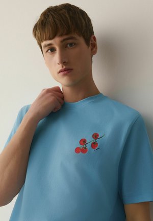Young man in a light blue shirt with a small tomato vine graphic on the chest, standing against a plain light wall.