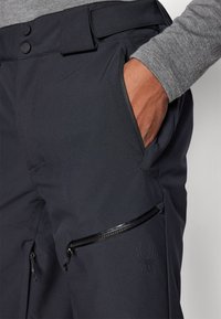 Pantalones técnicos negros con una textura suave, que cuentan con bolsillos con cremallera y un sutil logo de araña en el muslo. Cierre de dos botones en la cintura.