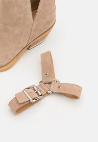 Botín de ante beige con tacón apilado, presenta un diseño recortado y una correa desmontable con hebilla metálica y detalles.