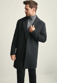 Mężczyzna poprawiający ciemnoszary płaszcz noszony na szarym swetrze z golfem i czarnych spodniach, patrzący w lewo na jednolity jasny tle.