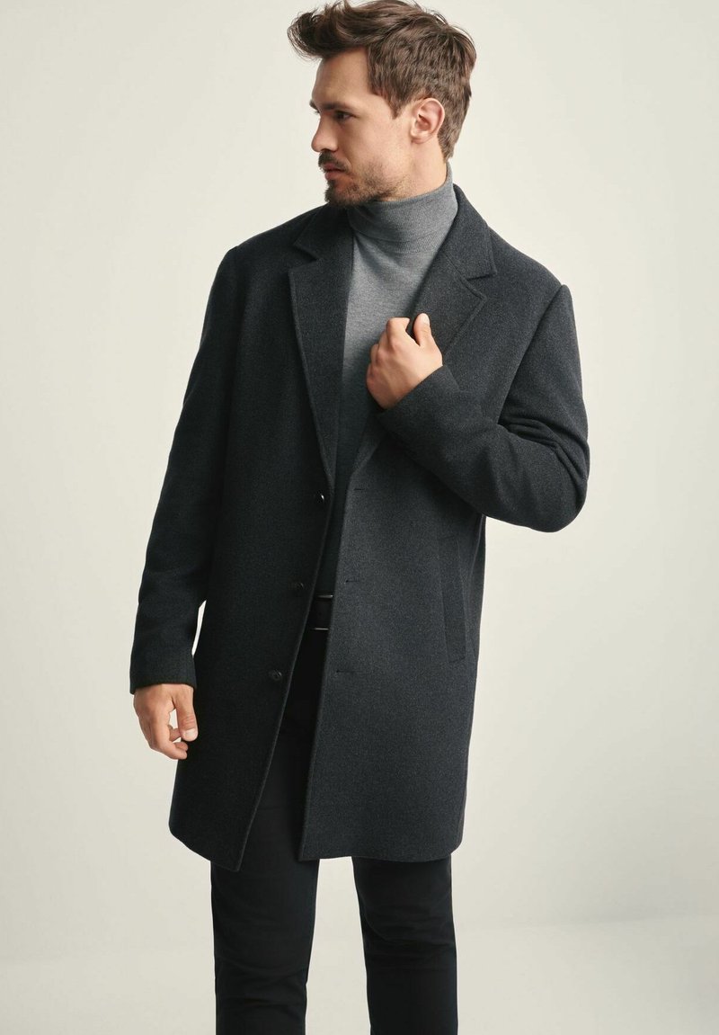 Mężczyzna poprawiający ciemnoszary płaszcz noszony na szarym swetrze z golfem i czarnych spodniach, patrzący w lewo na jednolity jasny tle.