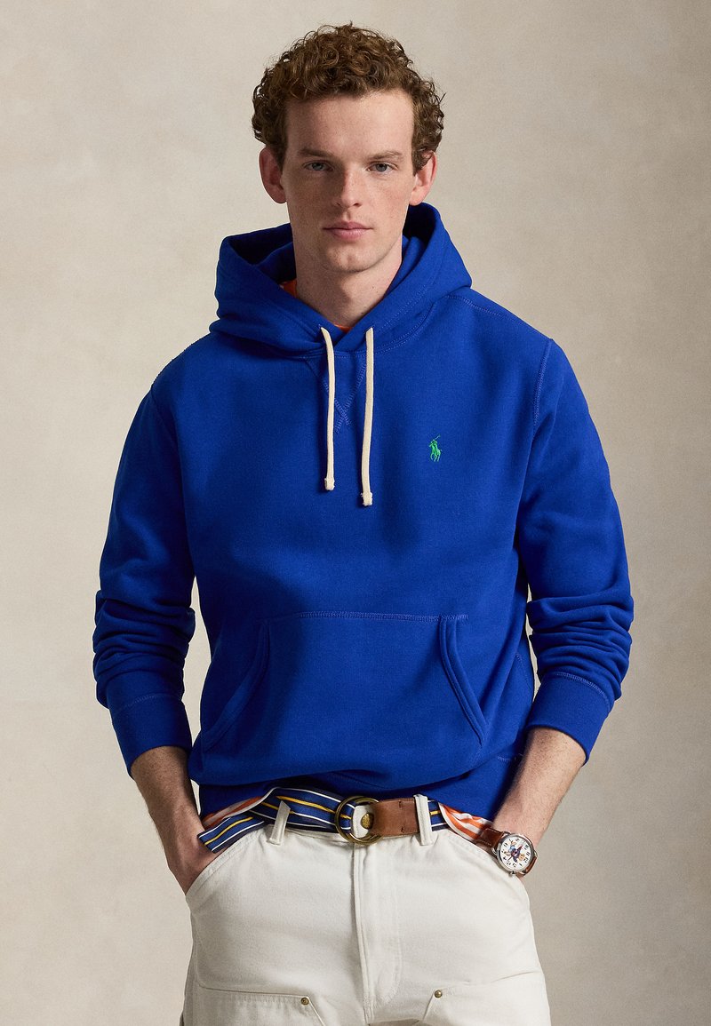 Jeune homme aux cheveux bouclés portant un sweat à capuche bleu vif, une ceinture rayée, un pantalon blanc et une montre, debout les mains dans les poches devant un fond uni.