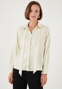 Blusa de satén color crema con diseño de botones, que presenta un cuello puntiagudo y cintas de tela en el frente, mangas largas con puños abotonados.