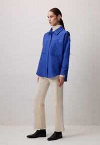 Camicia blu trapuntata con chiusura a bottone, due tasche frontali, indossata sopra un dolcevita beige e abbinata a jeans beige a zampa d'elefante e stivali neri.