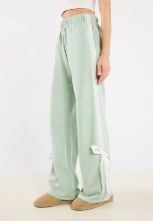 Luźne jasnooliwkowe spodnie dresowe z białymi bocznymi paskami i kokardkami przy mankietach, noszone z beżowymi butami wsuwanymi i białym krótkim topem.