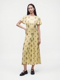 Femme portant une robe midi jaune à manches courtes à motifs et des chaussures noires à boucle, debout avec une main sur la hanche sur fond blanc.