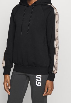 Sweat à capuche noir avec des bandes beiges marquées sur les manches et des leggings foncés assortis avec des lettres blanches sur la cuisse.