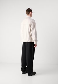 Giacca oversized di colore crema chiaro con colletto, orlo elasticizzato e maniche lunghe, abbinata a pantaloni neri larghi e scarpe nere.