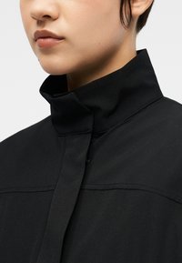 Artimas asmuo dėvi juodą striukę su aukšta apykakle ir slėptu priekiu, fone matomas vientisas fonas.