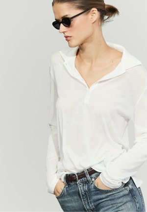 Kvinde med hår i knold iført sorte solbriller, en hvid skjorte med lange ærmer og åben krave samt blå jeans med hænderne i lommerne.