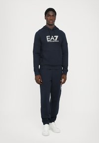 Felpa blu navy con logo bianco, tasca a marsupio e pantaloni della tuta coordinati con dettaglio del logo. Tessuto di cotone morbido, vestibilità casual e caviglie regolabili.