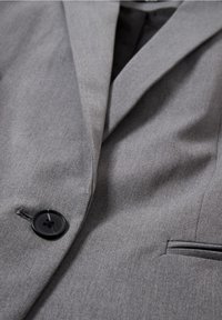 Gros plan d'une veste de costume grise montrant un bouton noir, un revers et une poche fendue sur un tissu lisse.