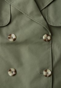Olivově zelený dvouřadý kabát z hladké látky, který má dvě velké kulaté knoflíky s mramorovým vzorem a výrazné ramenní klopy.
