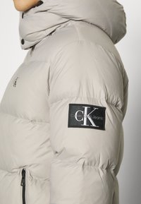 Beige pufferjack met capuchon en zwart Calvin Klein Jeans logo patch op de bovenarm, gedragen door een persoon die zijwaarts kijkt.