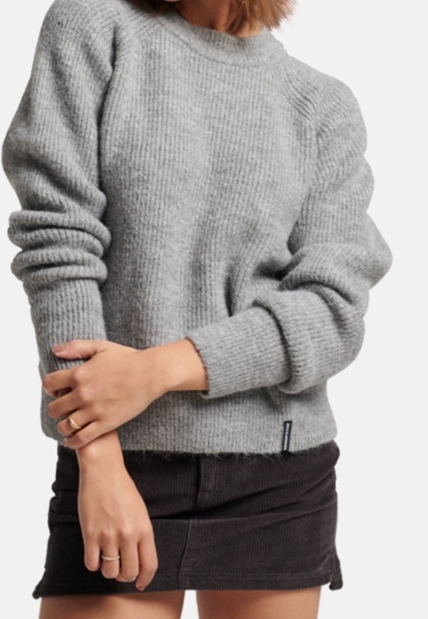 Personne portant un pull en maille gris à texture côtelée et une mini-jupe noire en velours côtelé, bras croisés devant.