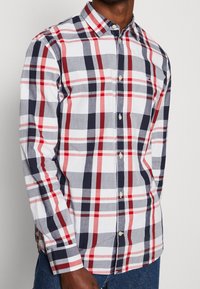 Camicia a quadri rossa, blu navy e bianca; colletto abbottonato; maniche lunghe; tessuto leggero; logo visibile sul petto a sinistra.