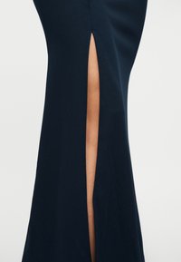 Κοντινό πλάνο μιας φούστας σκούρου ναυτικού μπλε με σκίσιμο μπροστά που αποκαλύπτει ένα γυμνό πόδι, πάνω σε απλό φωτεινό φόντο.