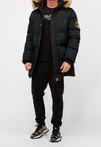 Doudoune noire avec capuche doublée de fourrure, arborant un badge sur la manche, portée par-dessus un t-shirt noir et un pantalon de survêtement noir, assortie à des baskets.