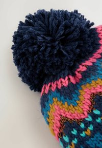 Bonnet en tricot bleu, rose, jaune et turquoise avec un grand pompon bleu marine sur le dessus ; présente des motifs géométriques et des textures variées.