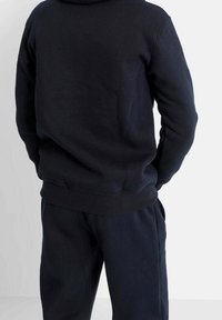 Sweat à capuche en polaire bleu marine avec une coupe décontractée et des poignets côtelés, porté par une personne vue de dos, assorti à un pantalon de jogging coordonné.