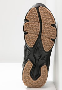 Semelle de chaussure de sport noire avec une bande de roulement en caoutchouc beige présentant des motifs en losange et géométriques, positionnée verticalement contre une surface blanche.