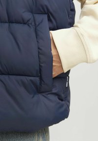 Jūras spēku izklāta veste ar šuvju tekstūru, ar sānu kabatu, uzvilkta virs gaiša toņa džemperis un džinsi. Minimāla zīmola redzamība.