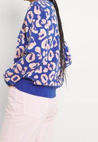 Pull bleu en tricot avec un motif léopard rose et bleu clair. Présente un ourlet et des poignets en côtes bleus. Associé à un pantalon rose clair.