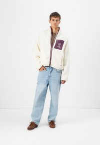White fleece jacket with a purple chest pocket, zipper closure, and side pocket. Paired with light blue denim jeans and brown shoes.