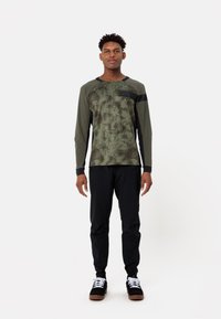 Chemise à manches longues vert olive avec un motif marbré, accents noirs et pantalon noir, accompagnée de baskets noires avec des lacets blancs.