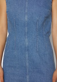 Vestido de mezclilla con cierre frontal, diseño ajustado y sutiles detalles en las costuras laterales. Color azul claro con una textura suave y forma estructurada.