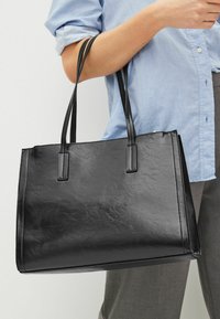 Bolsa tote de imitación cuero negra con dos asas resistentes. Forma rectangular, textura suave y brillo sutil. Combinando con una camisa de color azul claro.